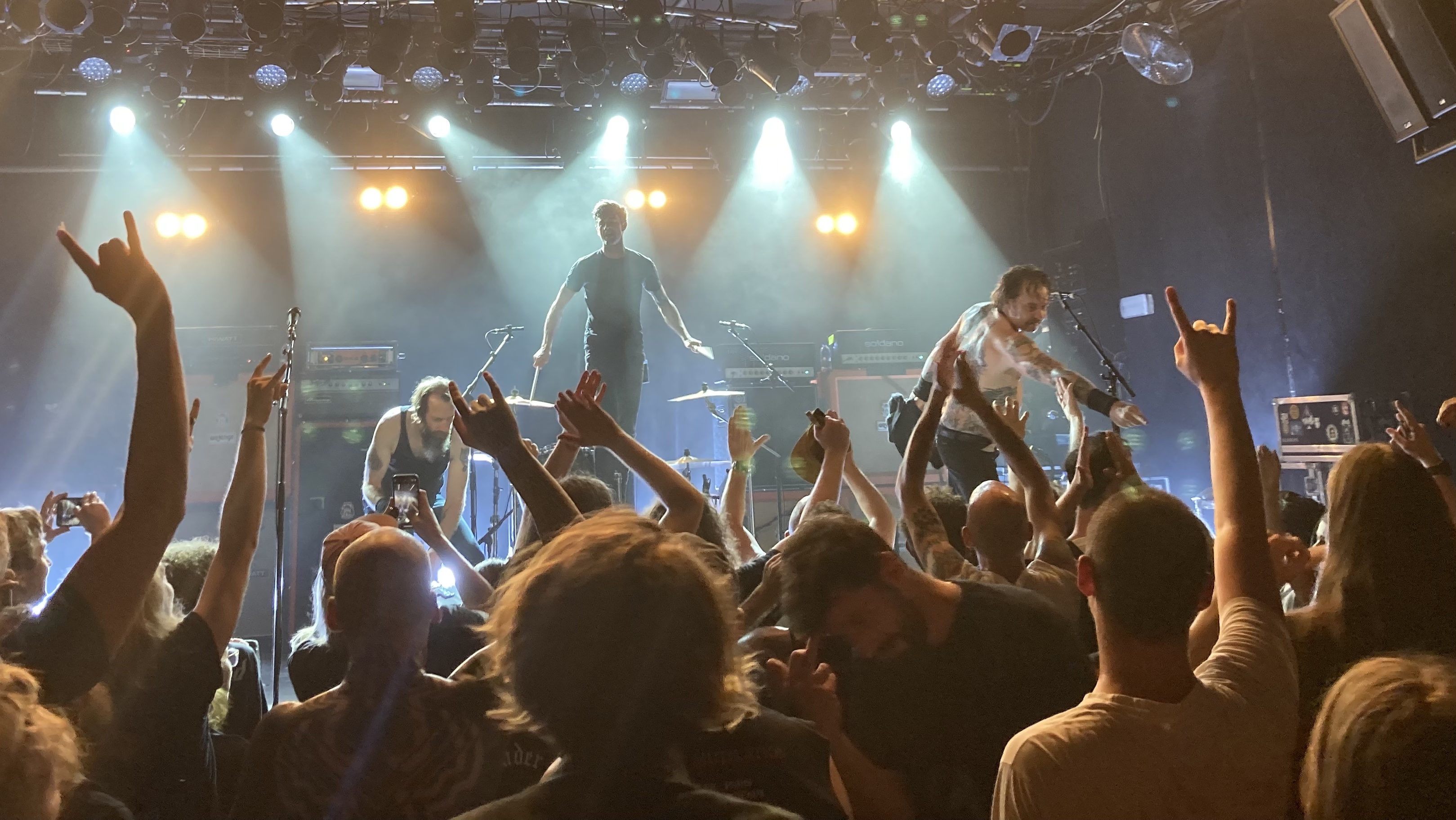 Photo of high on fire on stage at SO36, taken from a few meters away. The band is illuminated in white light. The drummer is standing on his seat, and the guitarist and bass player are interacting with the crowd. The crowd mostly has their hands up, many with metal horns.